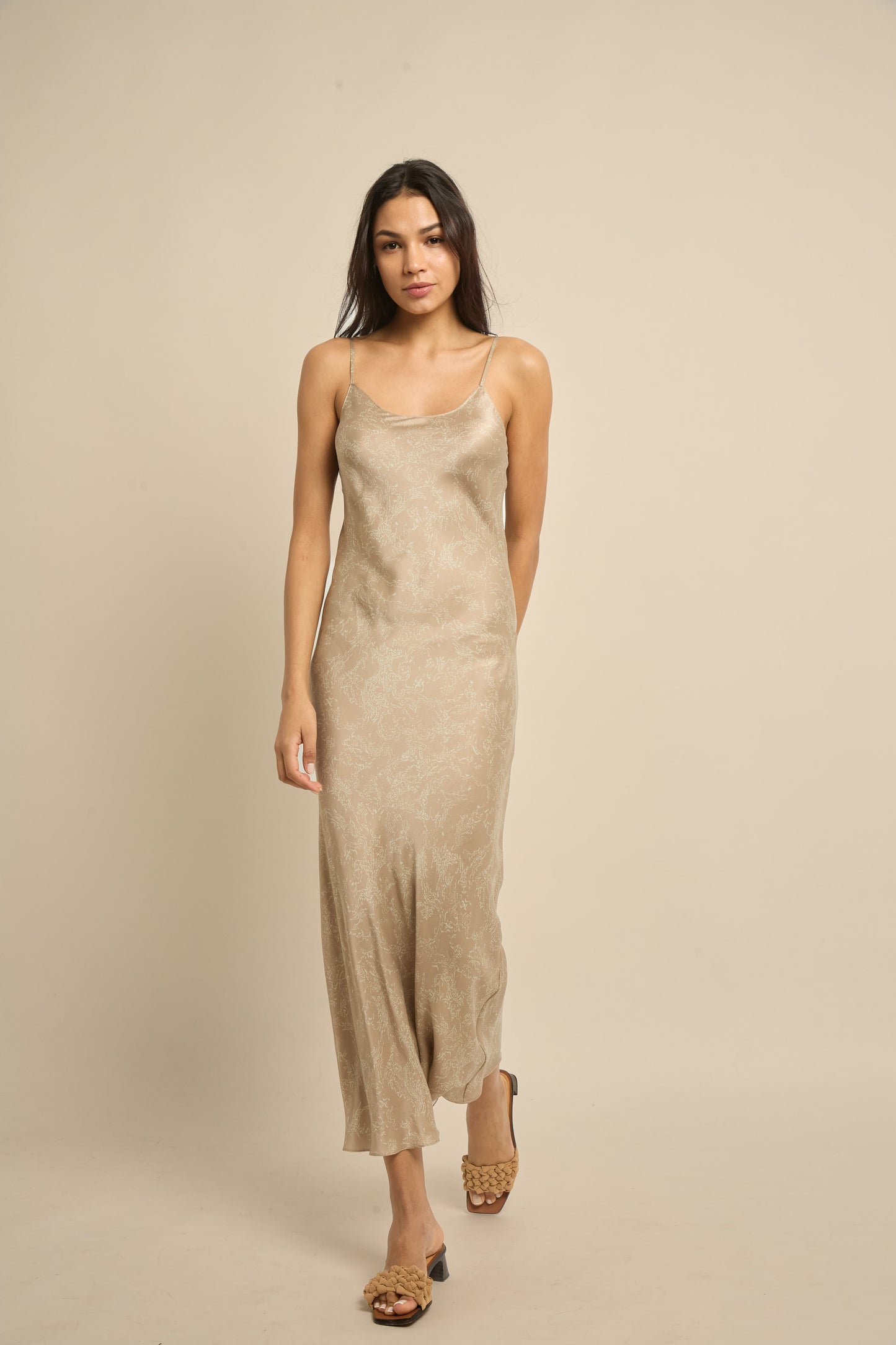 A woman wearing a beige midi silk slip dress with scoop neckline and narrow spaghetti straps.