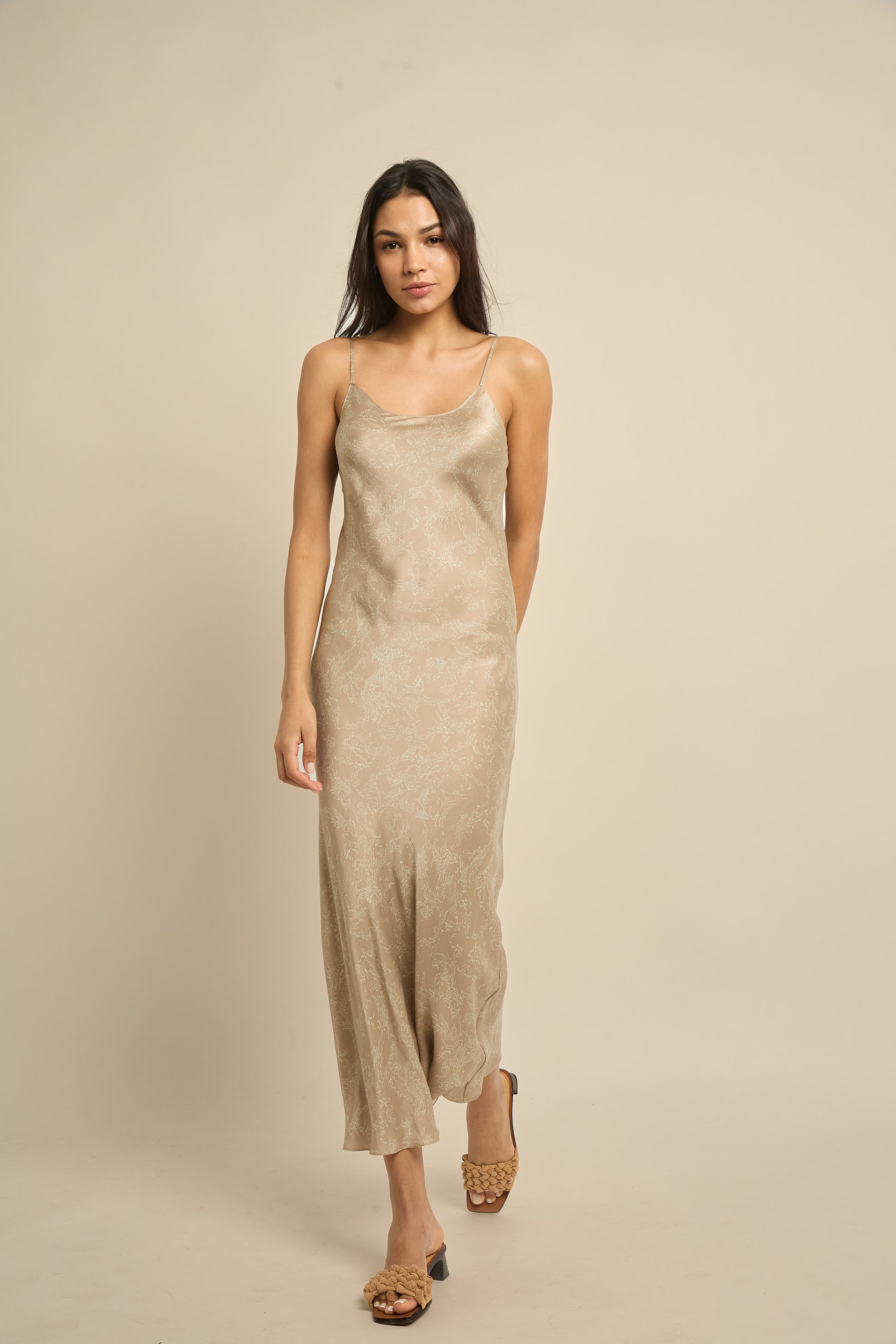 A woman wearing a beige midi silk slip dress with scoop neckline and narrow spaghetti straps.